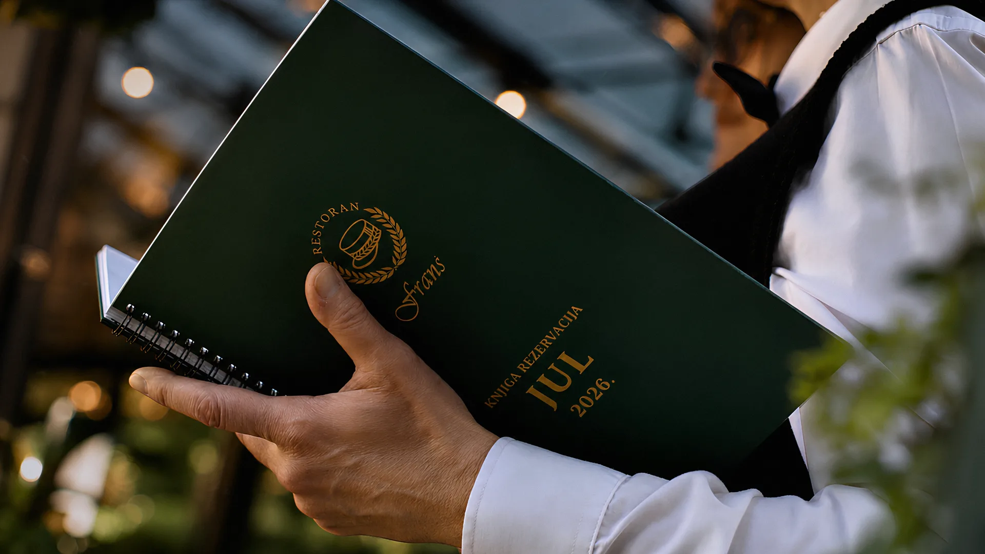 Waiter holding the green Franš reservation book for July 2026